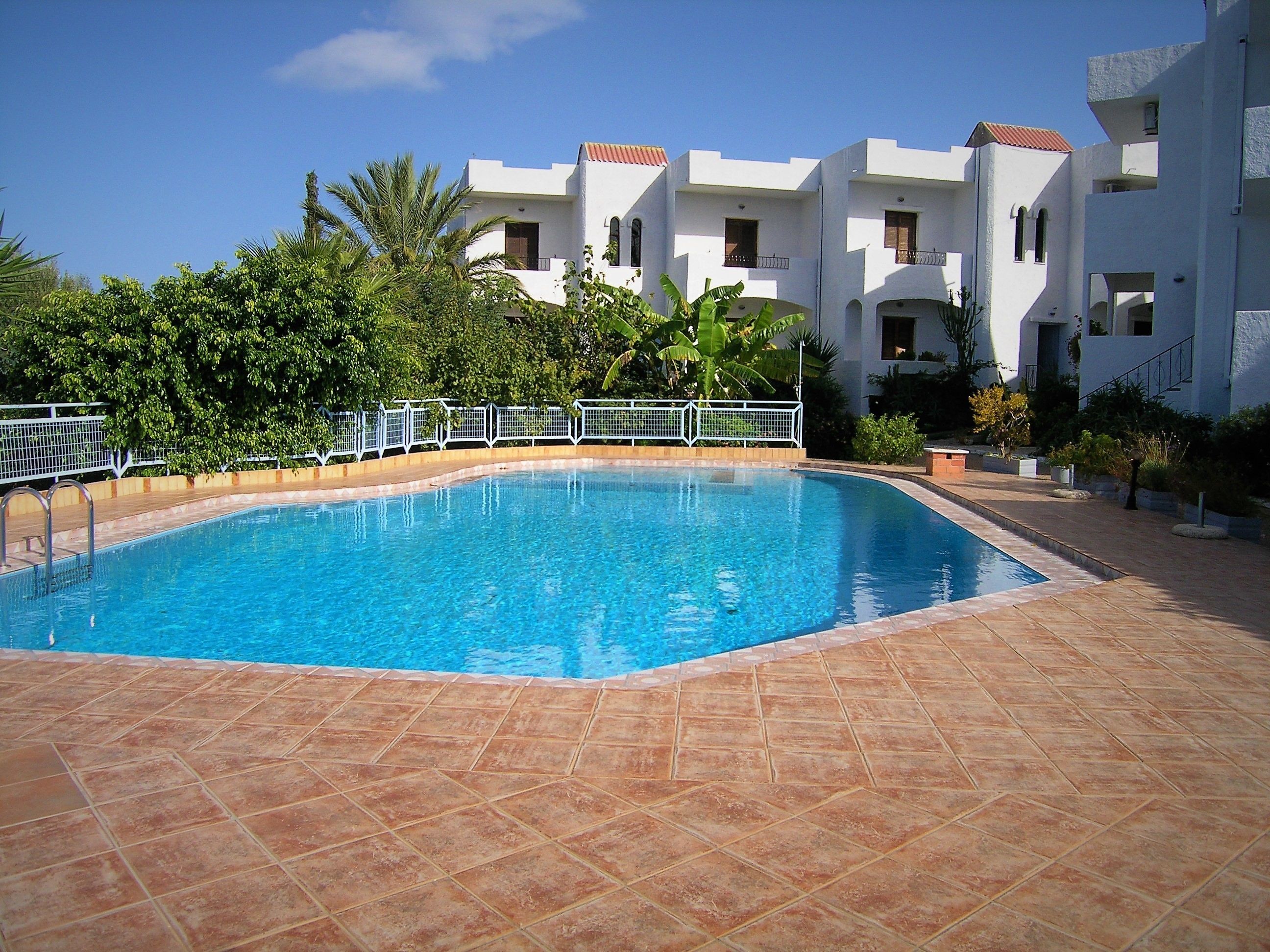 Una piscina rectangular con bordeado de baldosas, rodeada por una cerca blanca y abundante vegetación.