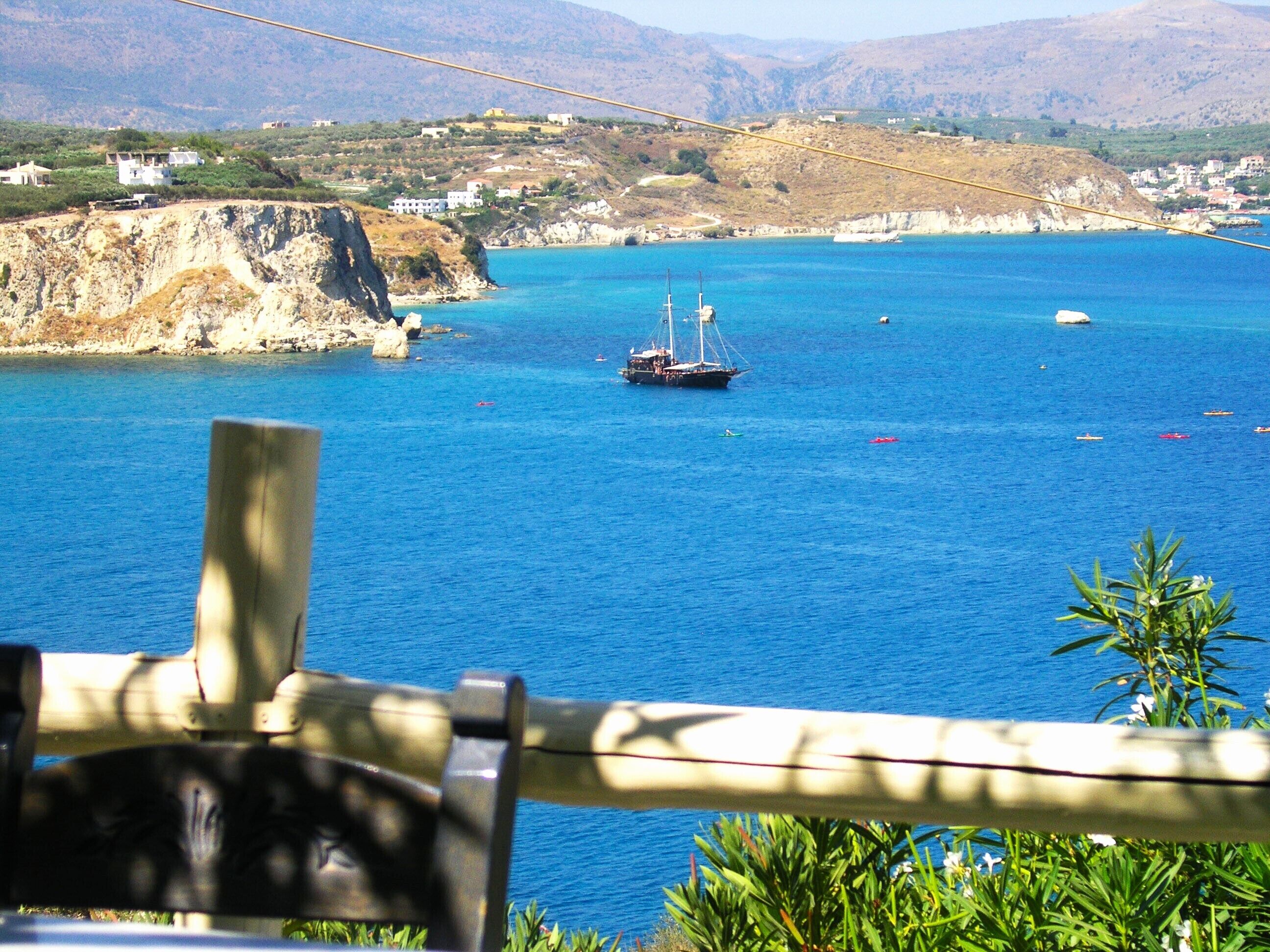 A coastal view with a wooden railing, a boat, and distant buildings on a hilly landscape.