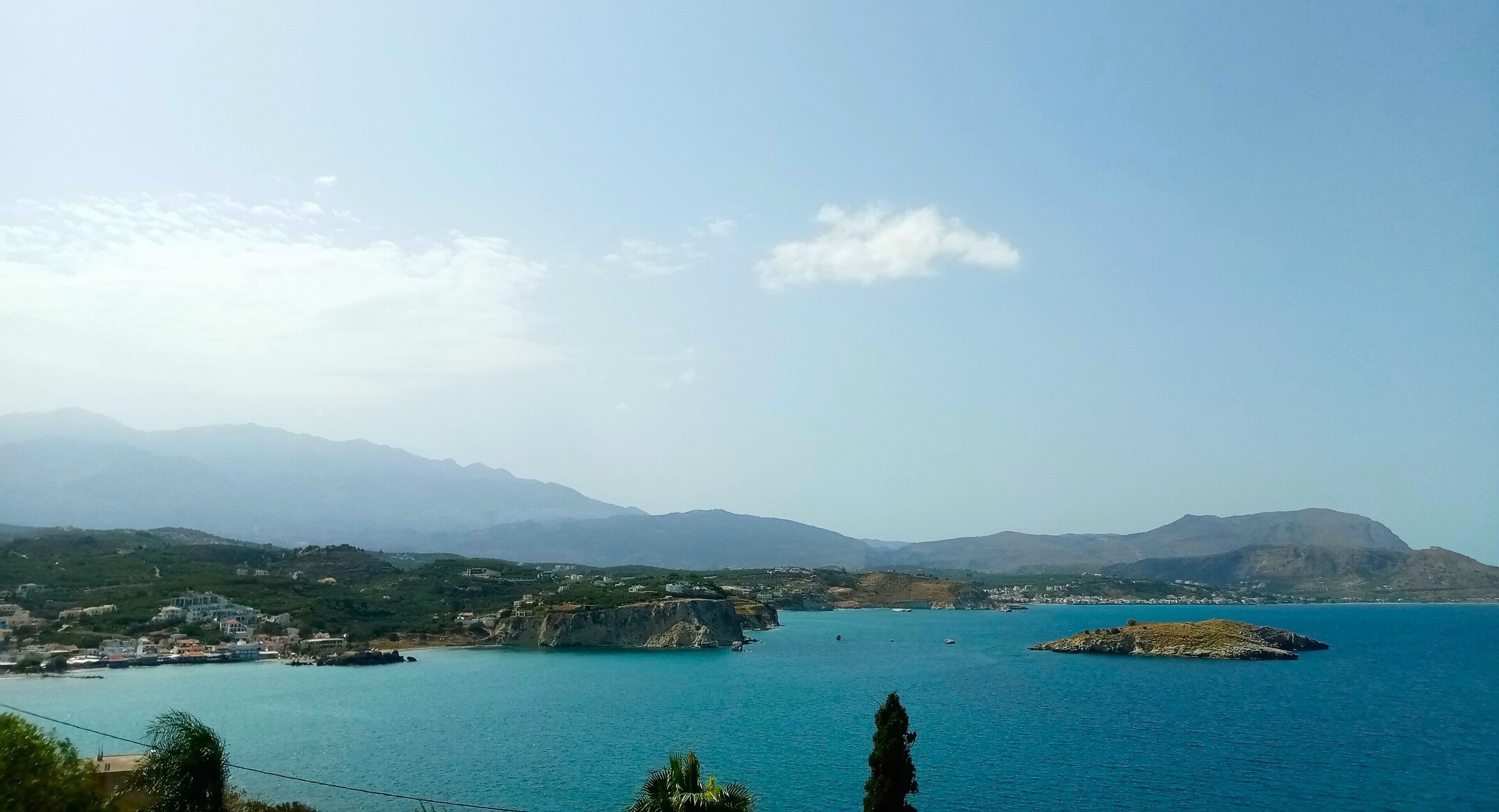 A coastal landscape with a clear view of the sea, a prominent rocky outcrop, and distant mountains.