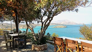An outdoor seating area with stone flooring, wooden tables, and chairs, overlooking a body of water and distant land. - Emerald (Apokoronas)