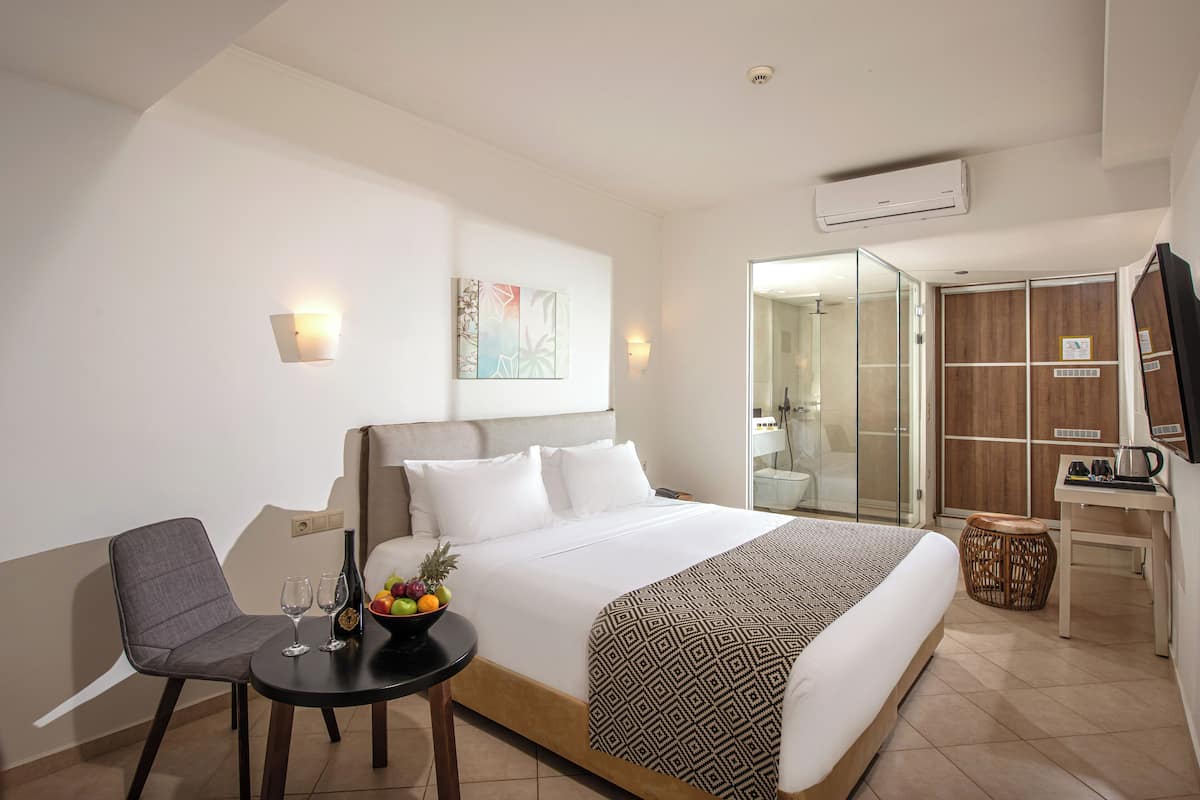 A hotel room with a large bed, a small table with a fruit bowl and glasses, a chair, a wardrobe, and a bathroom visible through a glass door.