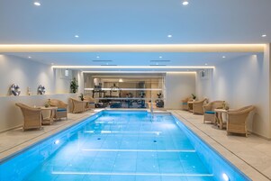 A modern indoor swimming pool with clear blue water, surrounded by wicker chairs and tables, and a reception area in the background.