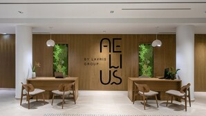 Una moderna zona de recepción con mostradores de madera, dos sillones, una pared verde con follaje y el logo 'AE LAVRIS GROUP'.
