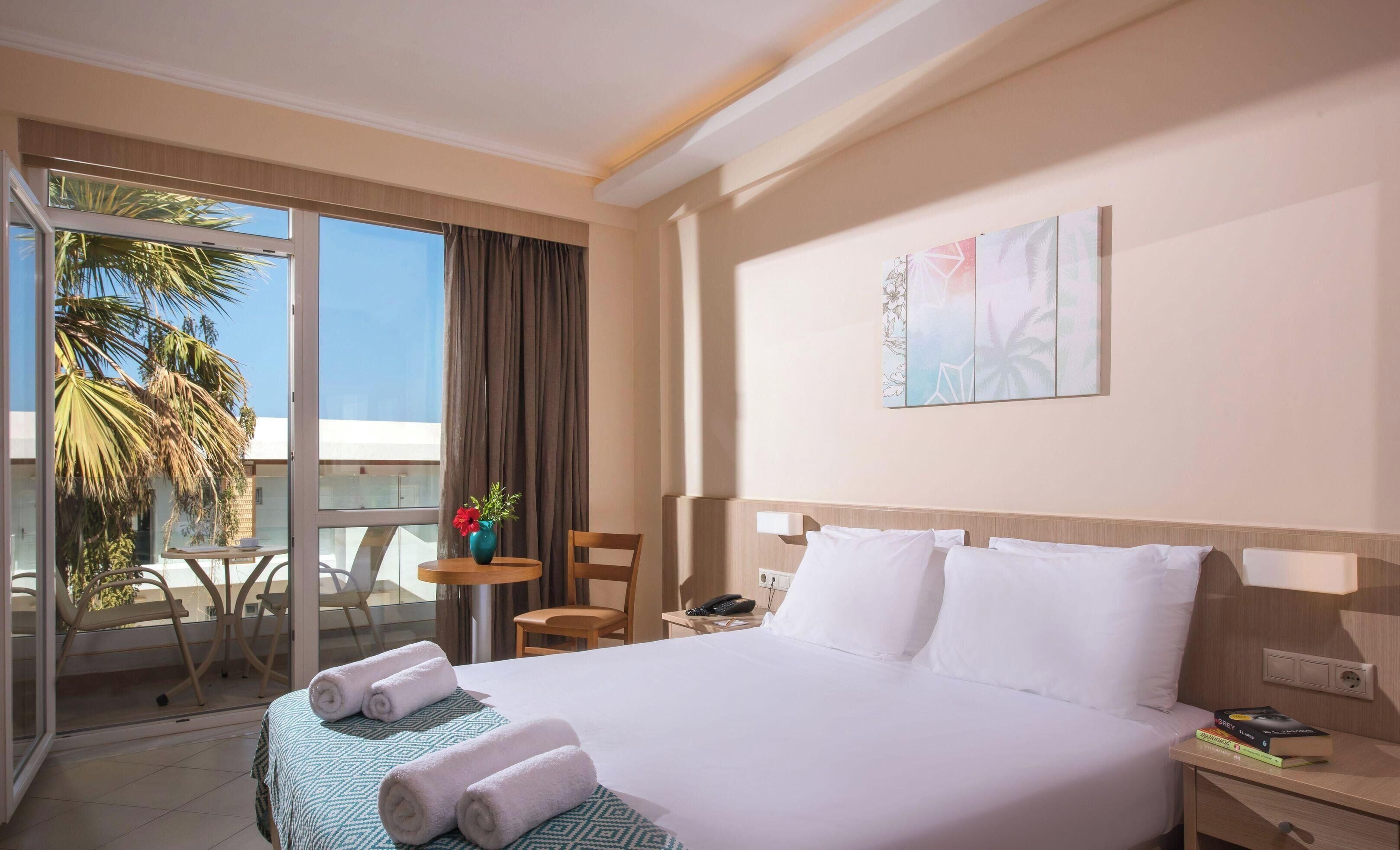 Habitación de hotel con cama, escritorio, silla, teléfono y ventana con vista a palmeras.