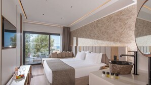 Una habitación de hotel moderna con una cama grande, una mesita de noche con frutero, un ventanal con vistas, un baño con bañera amplia y un espejo en la pared.
