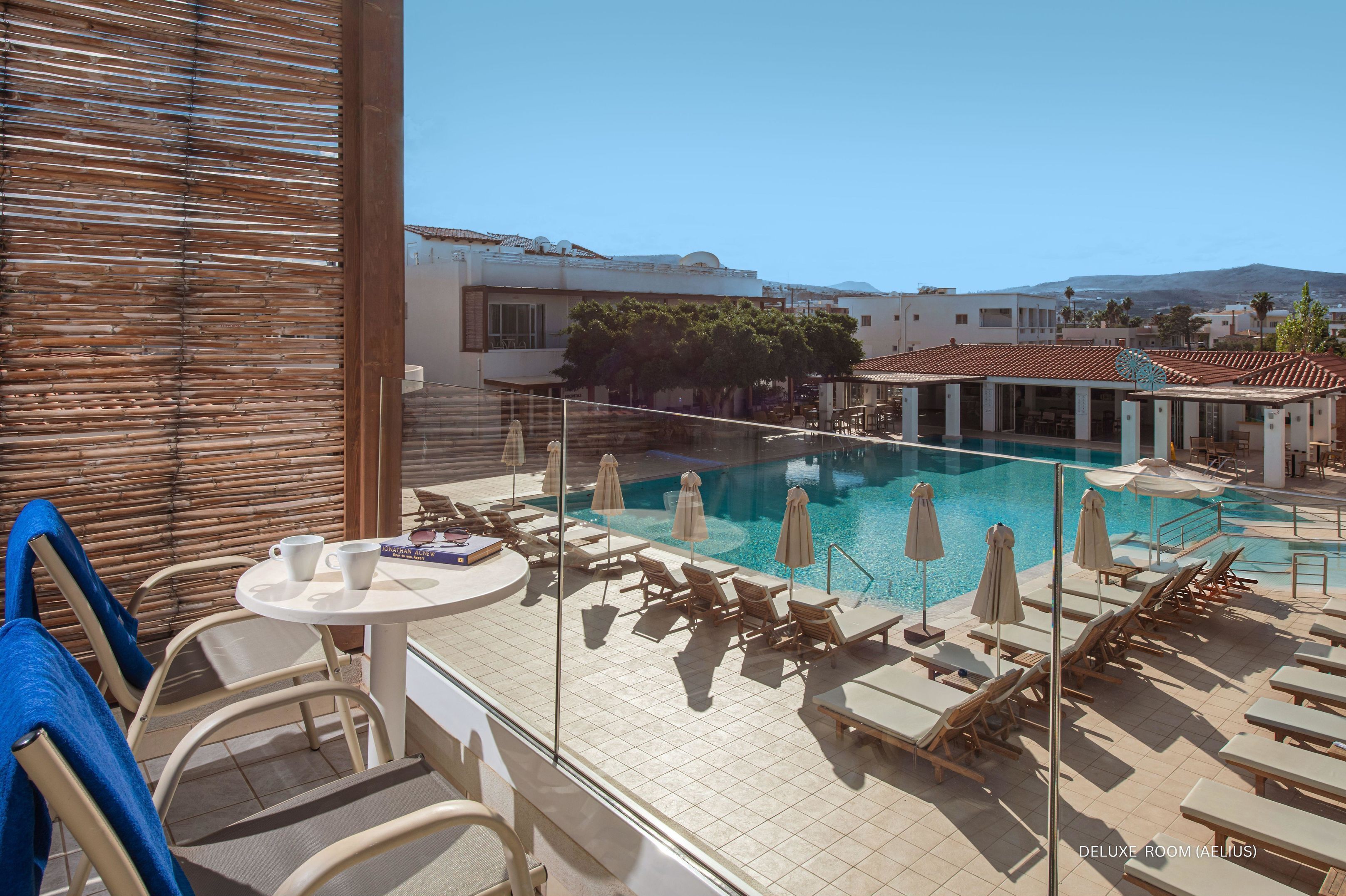 A pool area with lounge chairs and umbrellas, a table with a book and two cups, and a view of buildings and mountains in the background.