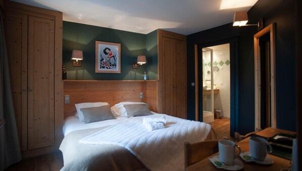 A neatly made bed with pillows, a bedside table with a cup and saucer, and a framed picture on the wall. - LOGIS HOTEL LE MONAL (Sainte-Foy-Tarentaise)