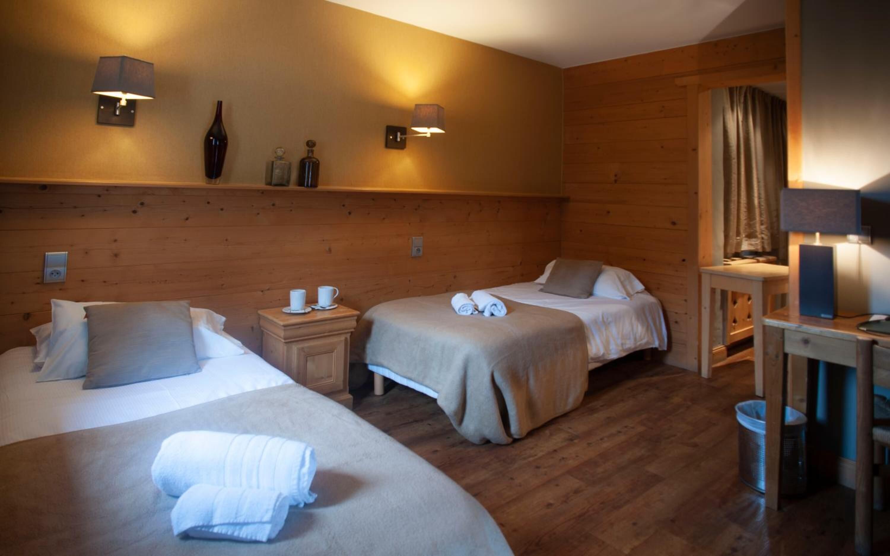 A hotel room with two beds, a nightstand, a small table, and a mirror. There are wall-mounted lamps and a shelf with decorative items.