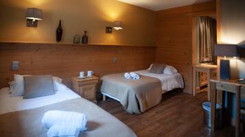 A hotel room with two beds, a nightstand, a small table, and a mirror. There are wall-mounted lamps and a shelf with decorative items.
