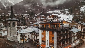 A town with a prominent tower, a hotel named 'HOTEL LE MONAL,' and snow-covered mountains in the background.