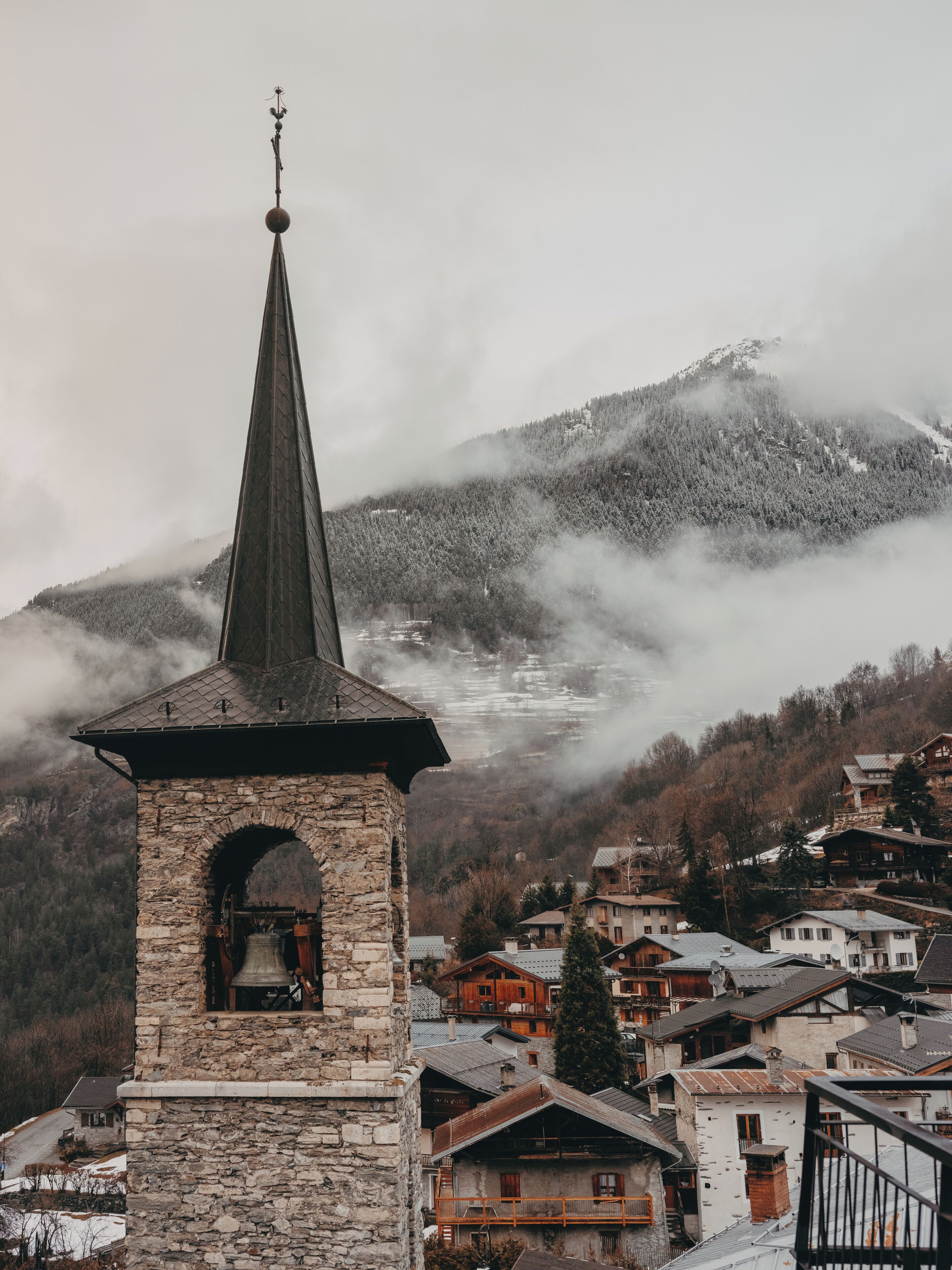 A stone bell tower with a spire, surrounded by traditional houses with sloped roofs, set against a backdrop of a snow-covered mountain and mist.