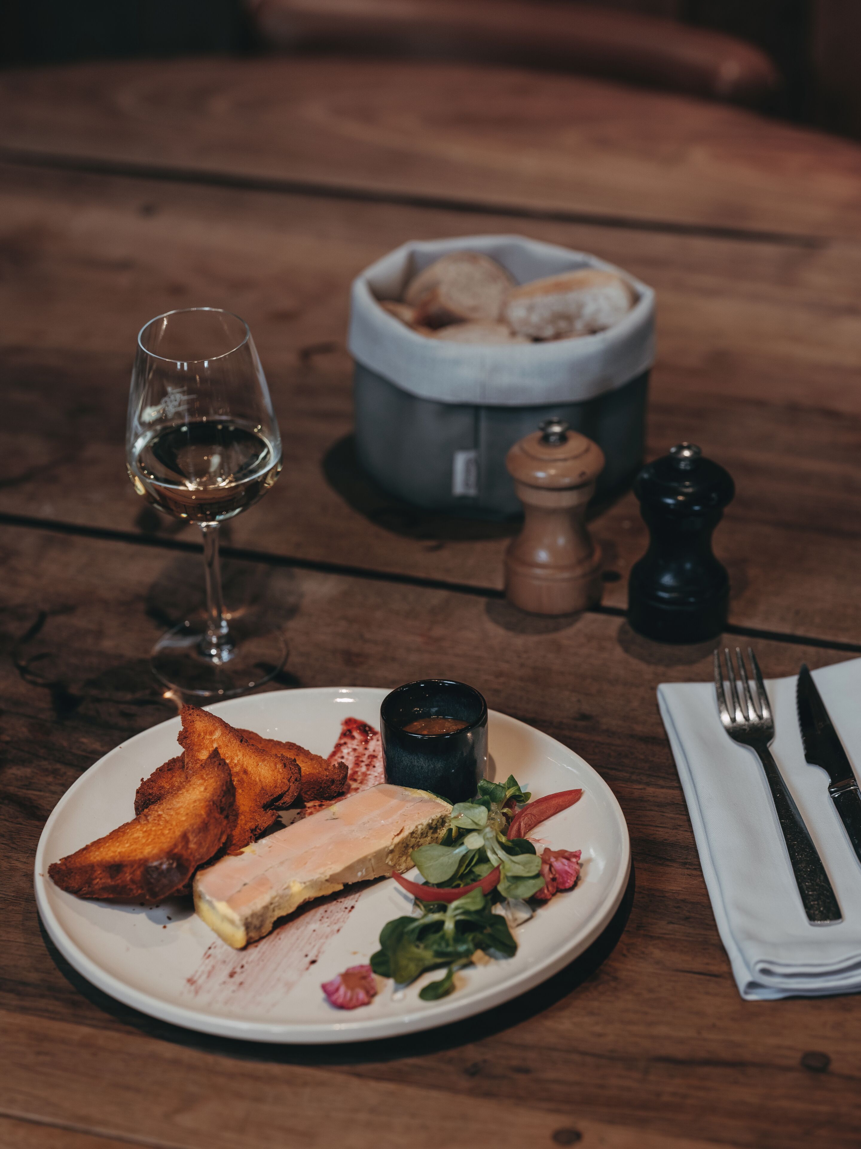 Een eettafel met een bord eten, een glas wijn, zout- en pepervaatjes, en een servet met bestek.