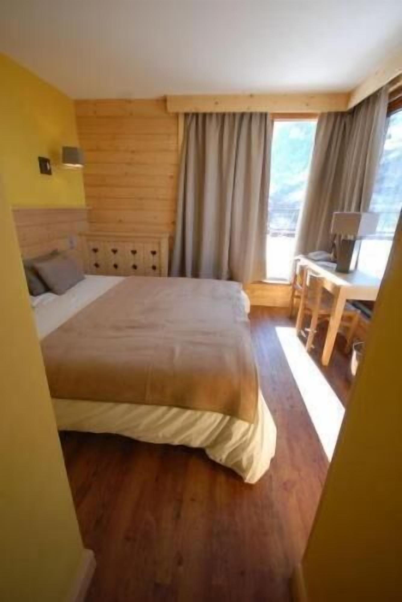 Een slaapkamer met een groot bed, een houten hoofdbord, een bureau met stoelen en een raam met gordijnen.