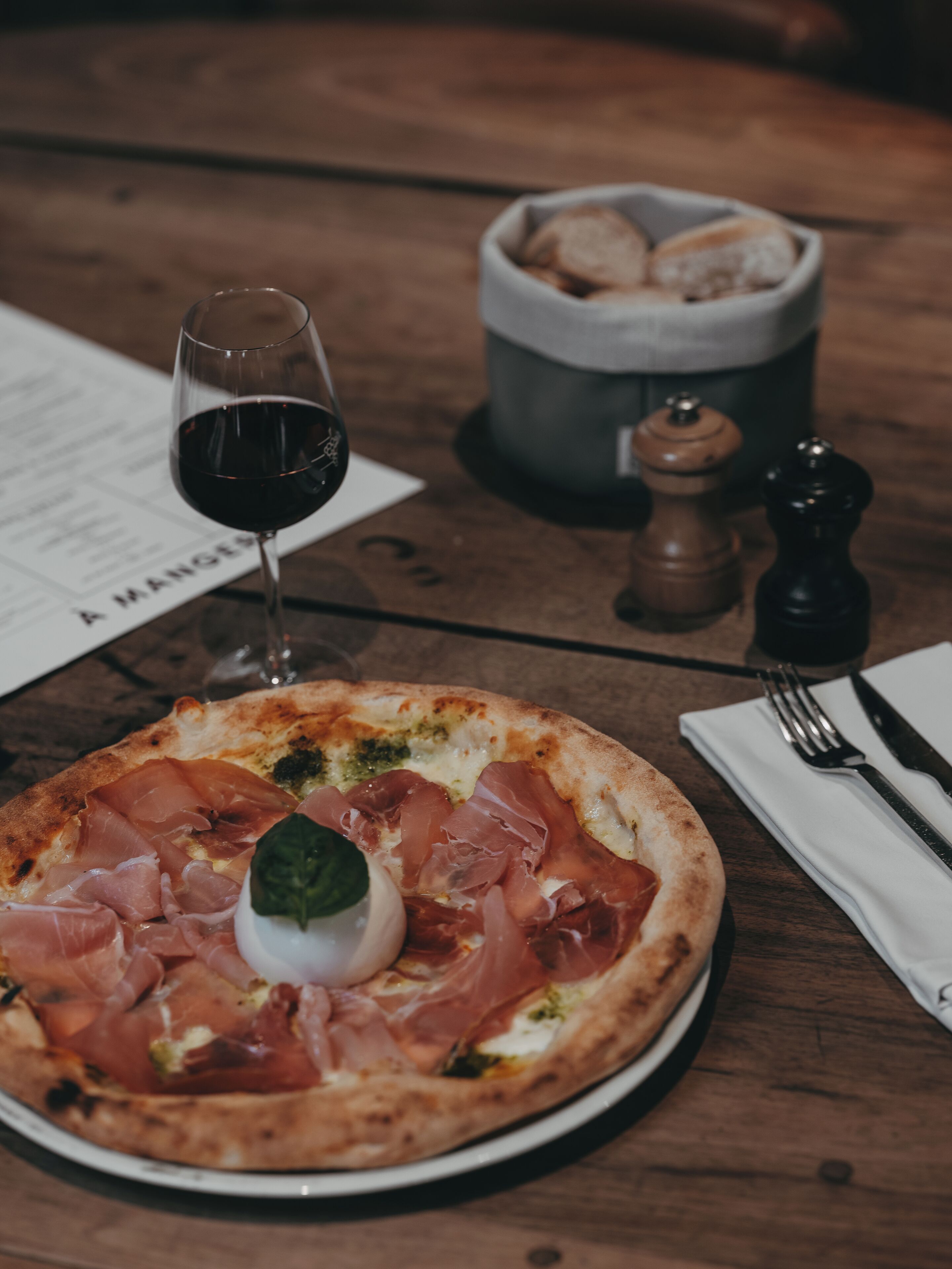 A pizza with prosciutto and a basil leaf on a plate, a glass of red wine, salt and pepper shakers, and a basket of bread on a wooden table.