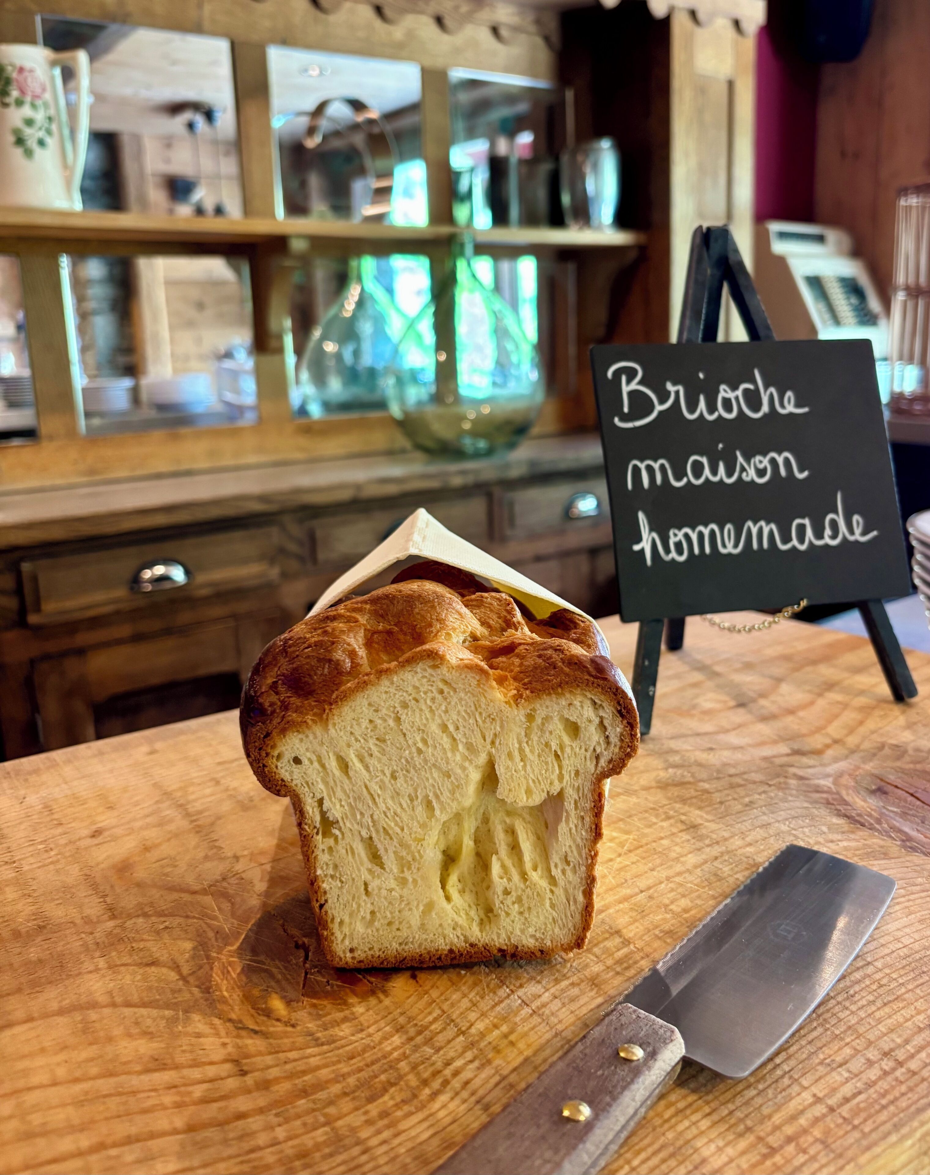 A loaf of brioche bread on a wooden cutting board with a knife beside it. In the background, there's a sign that reads 'Brioche maison homemade.'