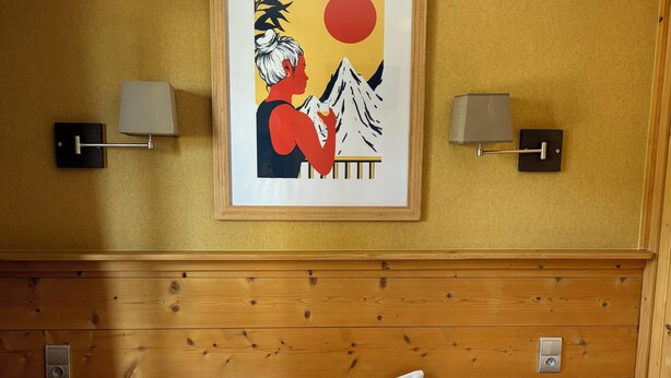 A hotel room with a wooden headboard, a bed with white linens, a framed artwork above the bed, and two wall-mounted lamps. - LOGIS HOTEL LE MONAL (Sainte-Foy-Tarentaise)