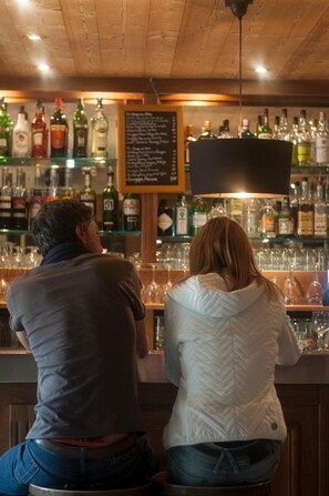 A bar with a wooden ceiling and a black hanging light fixture. There are multiple bottles and glasses on shelves behind the bar. - LOGIS HOTEL LE MONAL (Sainte-Foy-Tarentaise)