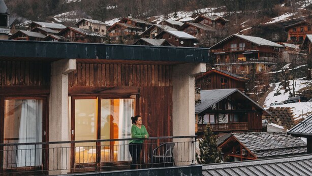 A mountain village with wooden houses, a balcony with a person, and snow-covered mountains in the background.