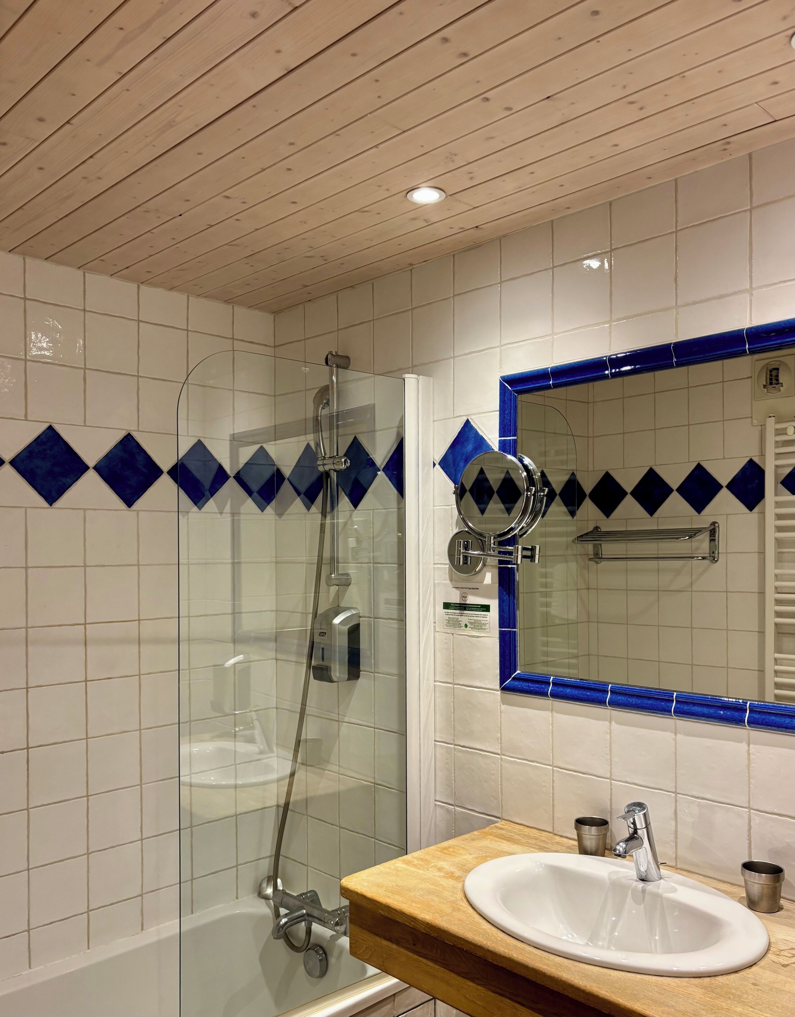 Un baño con ducha envuelta en vidrio, techo de madera, lavamanos blanco con encimera de madera y espejo con marco de azulejos azules.