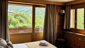 A hotel room with a bed, a wooden dresser, a lamp, and large windows offering a view of a forested area.