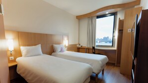 A hotel room with two beds, a desk, a window with curtains, and a view of the city. - ibis Barcelona Mollet (Mollet del Valles)