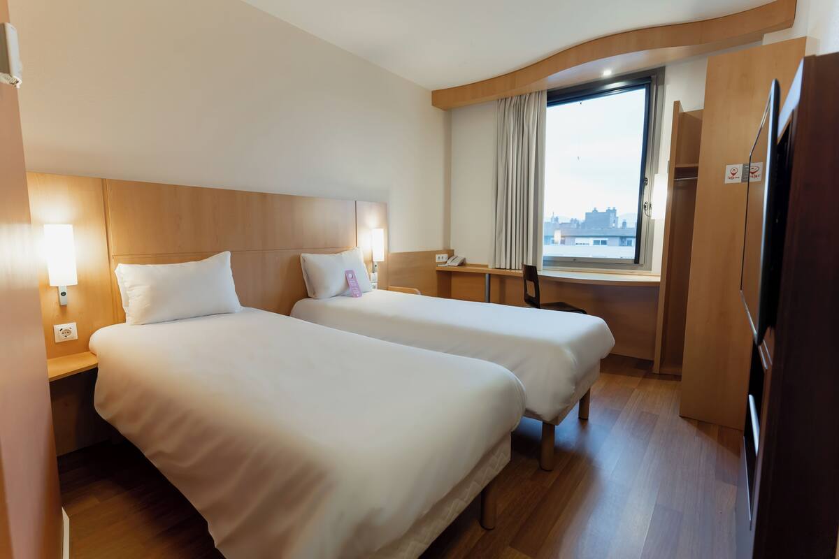 A hotel room with two beds, a desk, a window with curtains, and a view of the city.