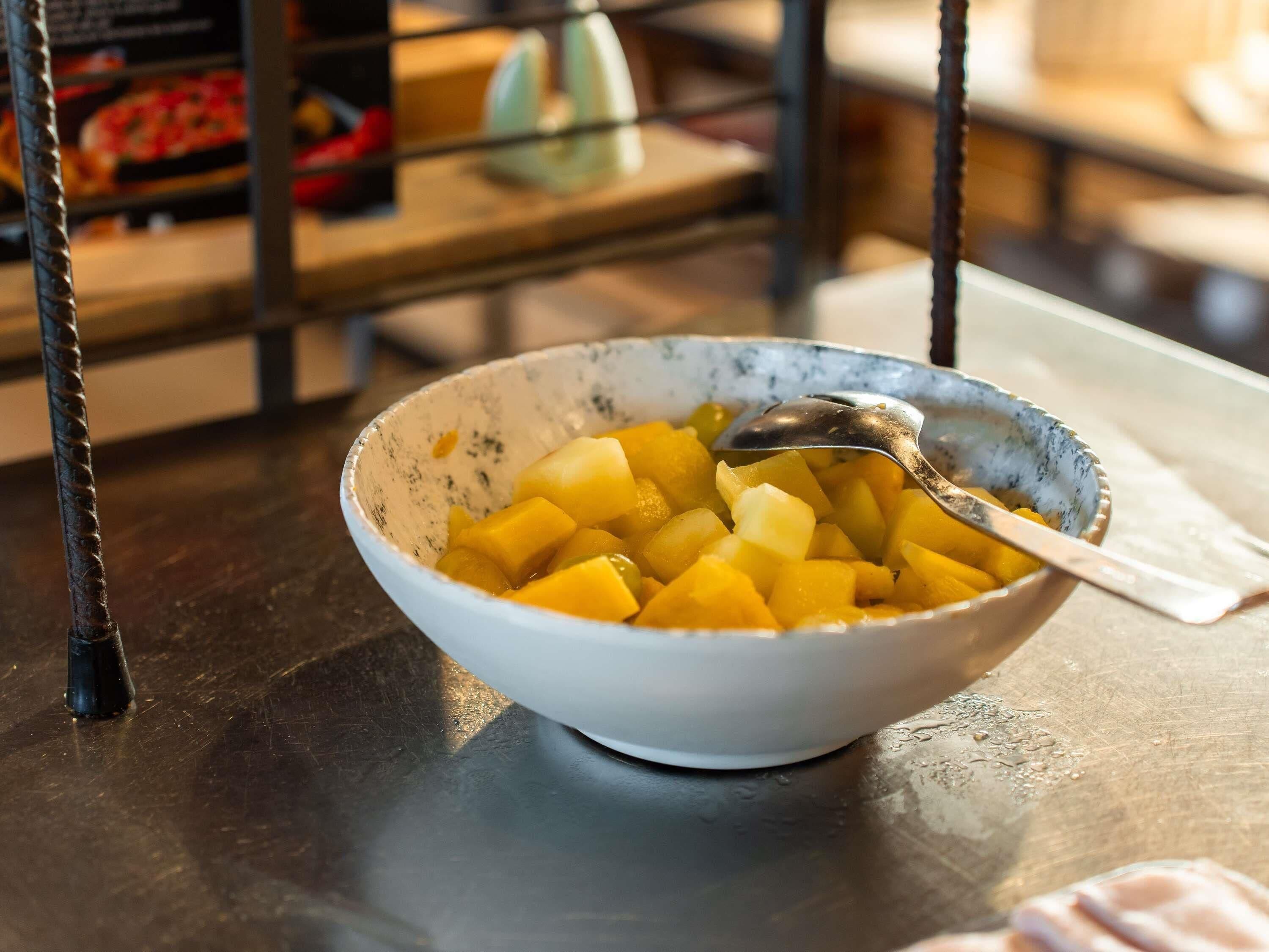 Eine Schüssel gewürfeltes Obst auf einem Tisch, zusammen mit einem Löffel, in einer Umgebung mit einem Metallgestell und unscharfem Hintergrund.