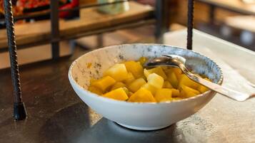 Eine Schüssel gewürfeltes Obst auf einem Tisch, zusammen mit einem Löffel, in einer Umgebung mit einem Metallgestell und unscharfem Hintergrund.