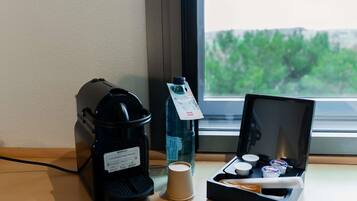 A coffee maker, a water bottle, a cup, and a box with condiments on a table by a window.