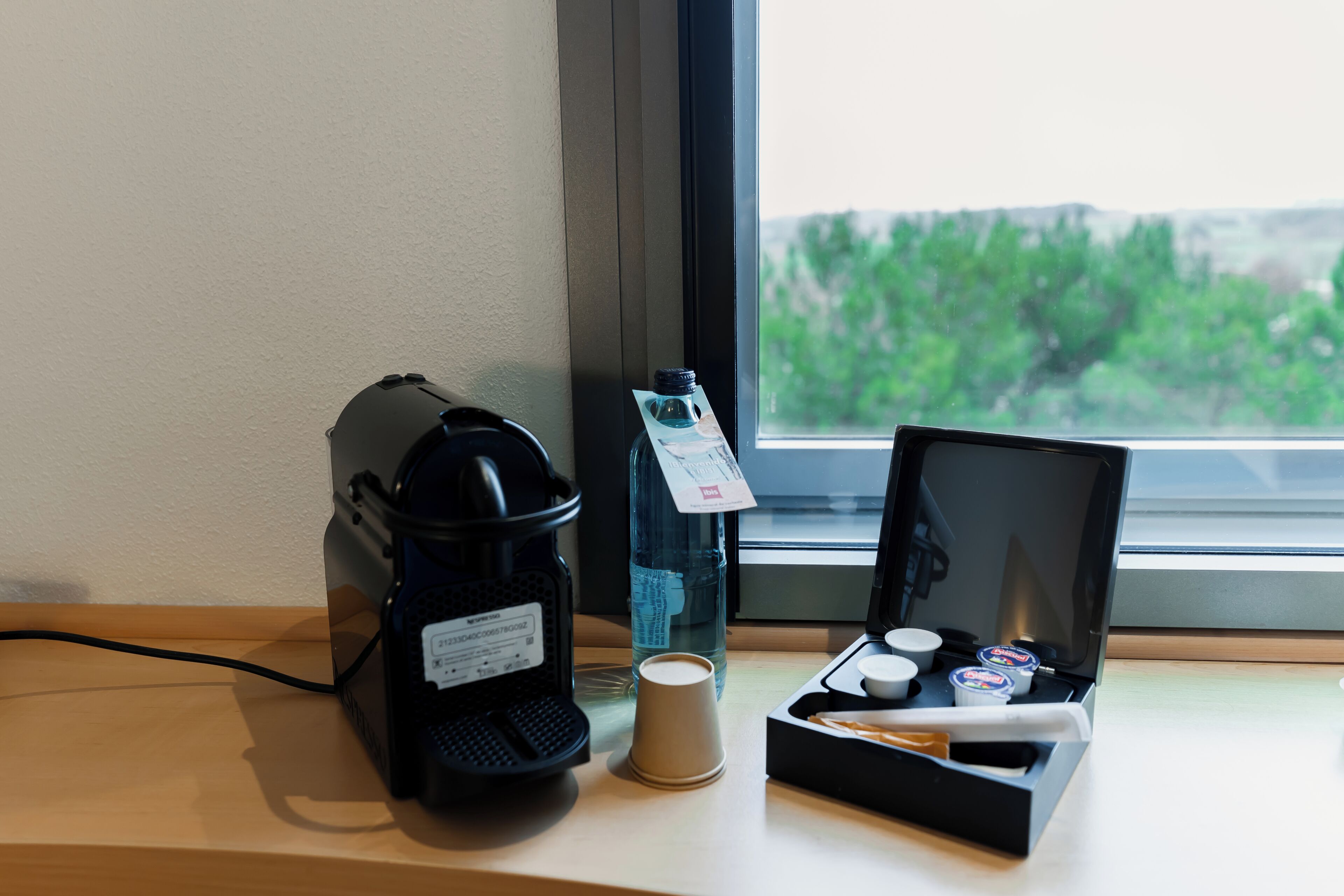A coffee maker, a water bottle, a cup, and a tray with condiments on a table near a window.
