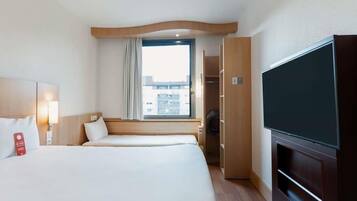 Ein Hotelzimmer mit zwei Betten, einem Fernseher, einem Kleiderschrank und einem Fenster mit Blick auf Gebäude.
