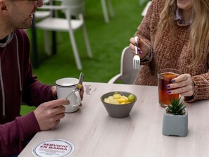 Dos personas sentadas en una mesa al aire libre, con un cuenco de fruta, una taza y un vaso de té.
