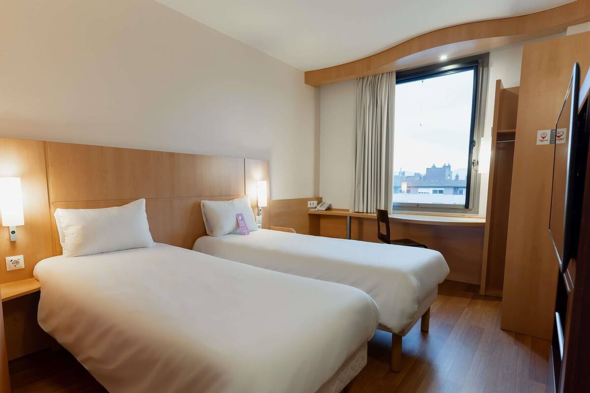 A hotel room with two beds, a desk, a chair, a window with curtains, and a city view.