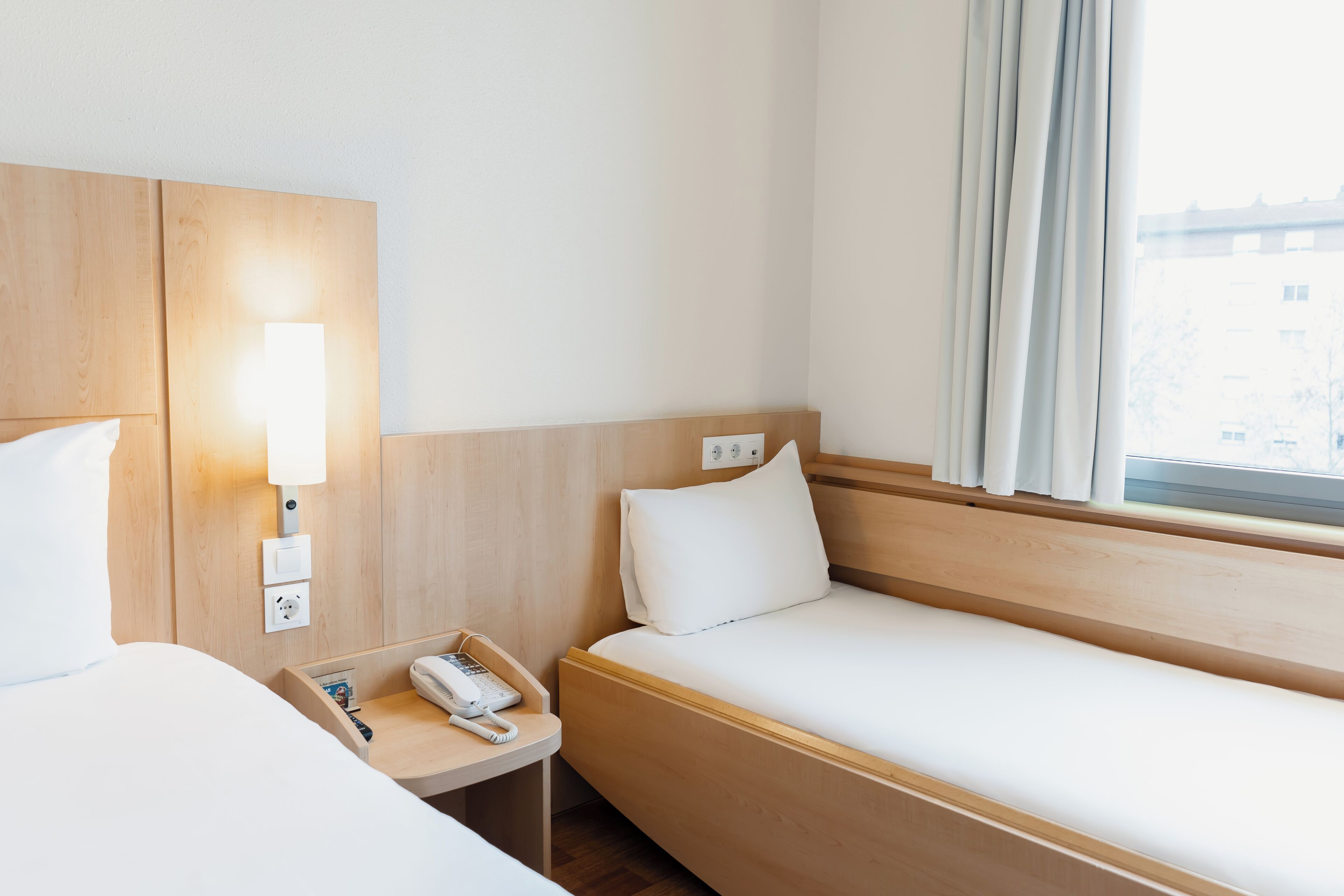 A hotel room with a wooden headboard, a bedside table with a phone, a lamp, and a window with curtains.