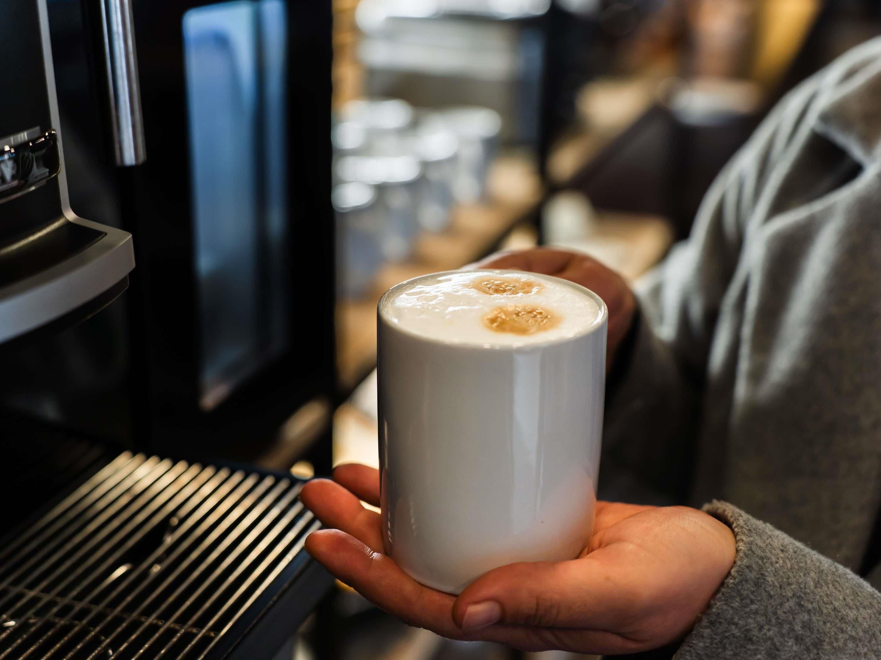 A hand holding a white mug with a frothy beverage, in front of a coffee machine.
