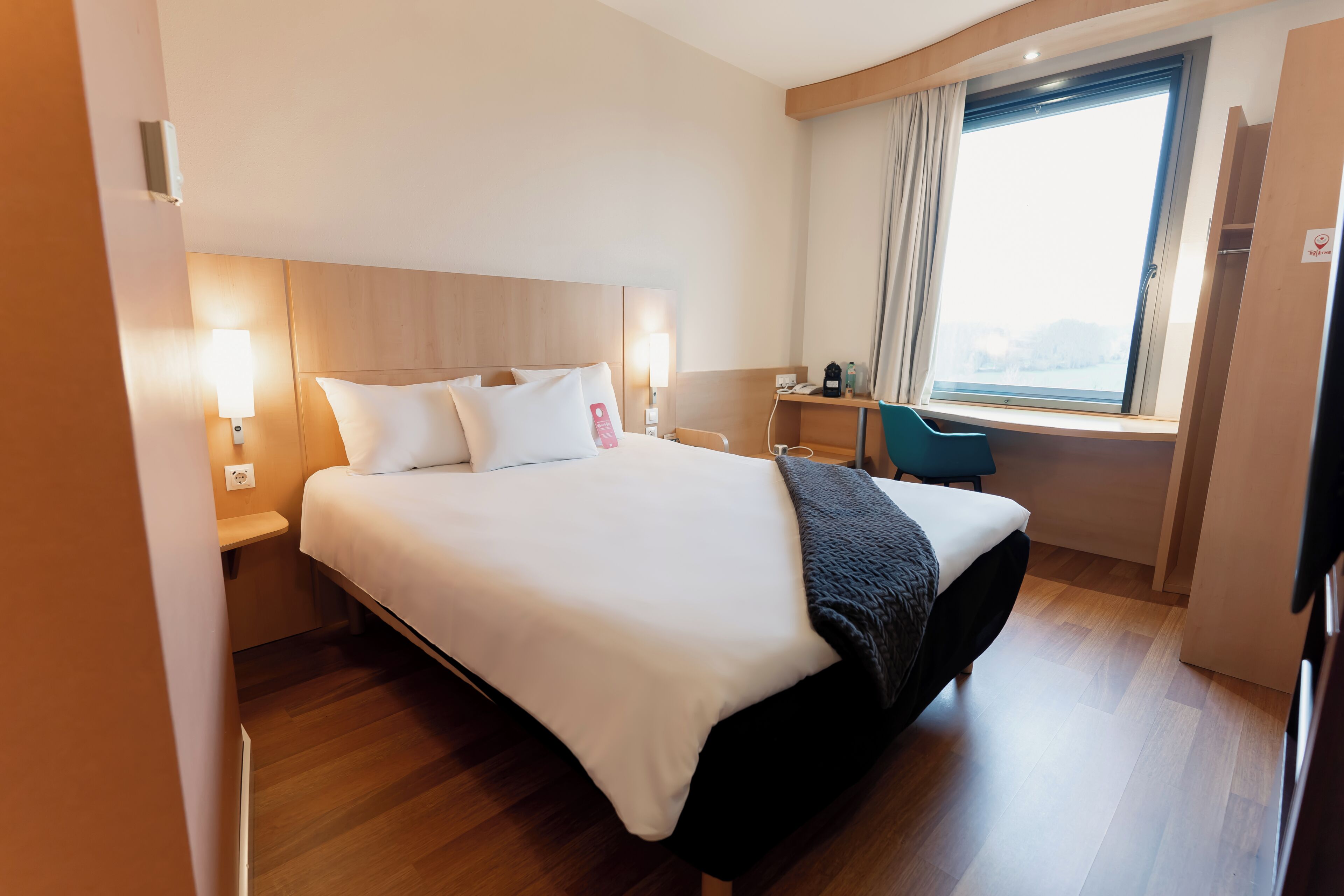 A hotel room with a large bed, bedside lamps, a desk, a chair, and a window with curtains.