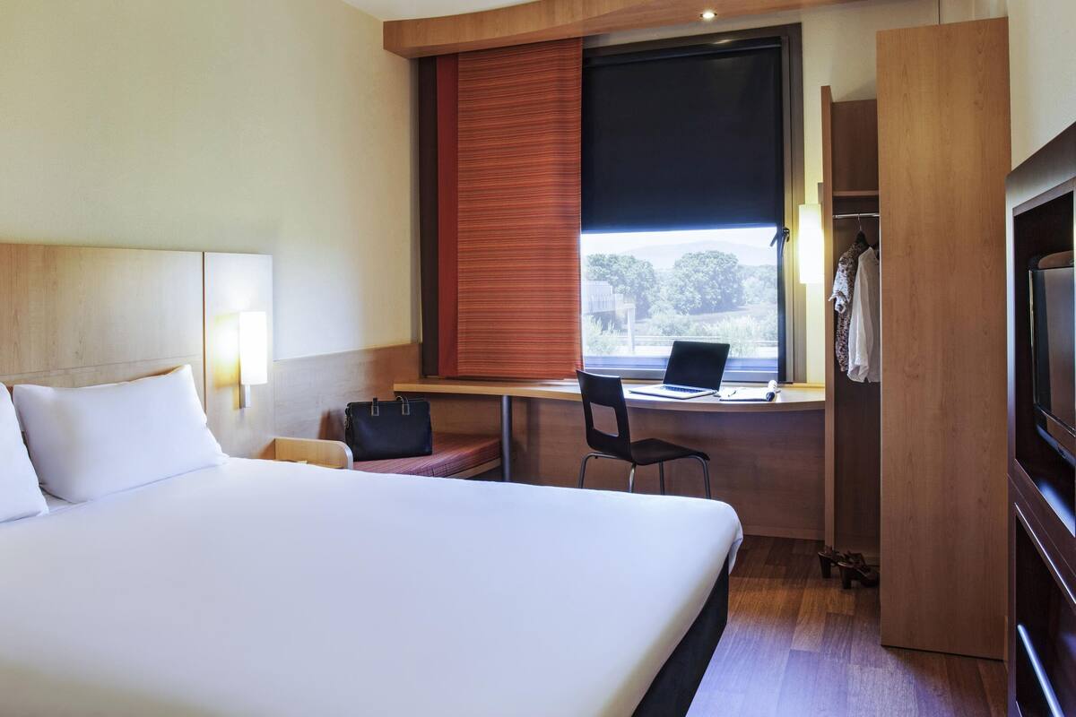 Une chambre d’hôtel comprenant un lit, un bureau avec un ordinateur portable, une chaise, une armoire et une fenêtre donnant sur des arbres.