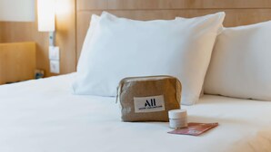 Una cama bien hecha con sábanas blancas, una bolsa de yute con etiqueta, un frasco pequeño y una tarjeta sobre la cama.
