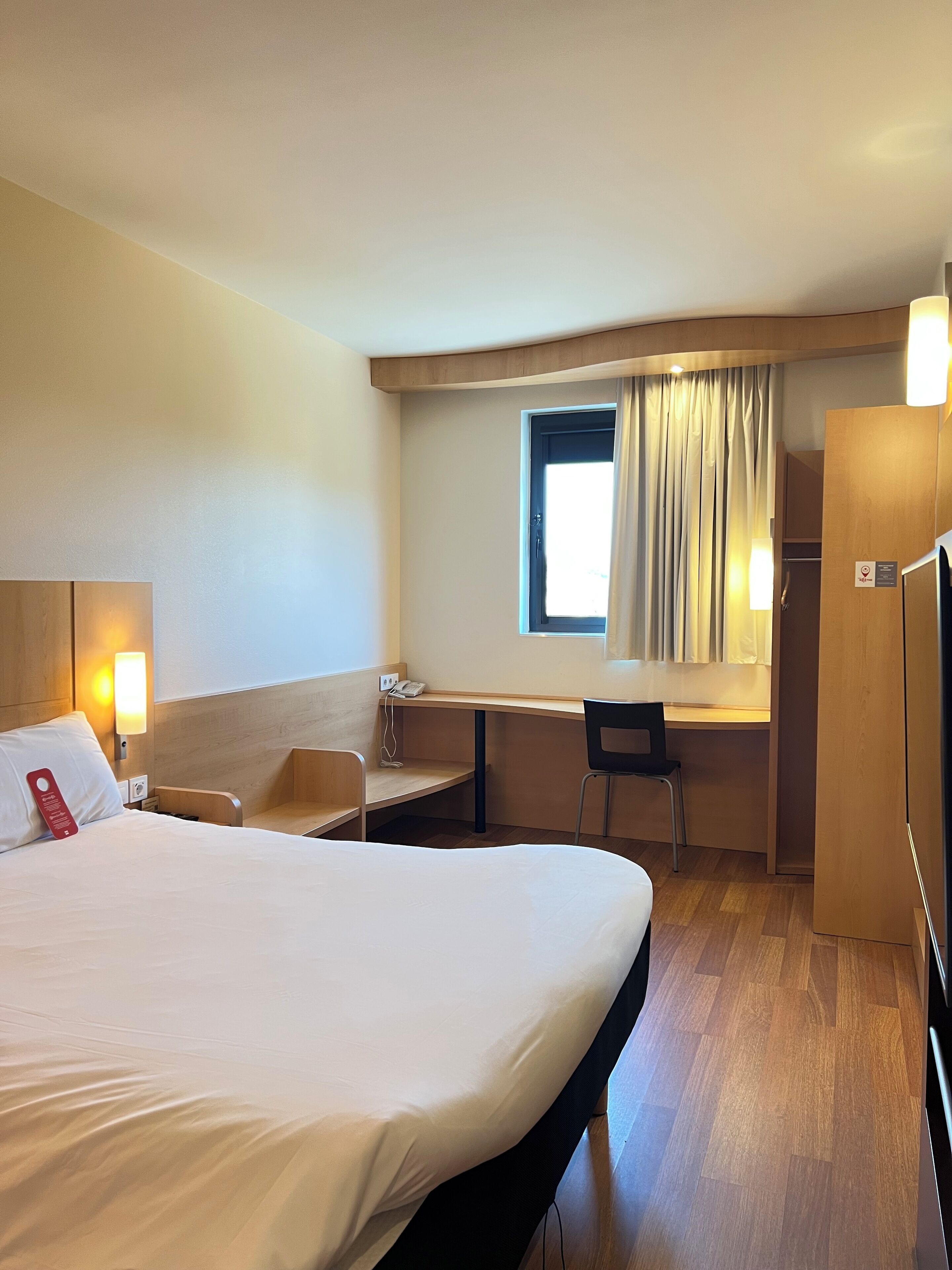 A hotel room with a bed, desk, chair, and a small window with curtains.