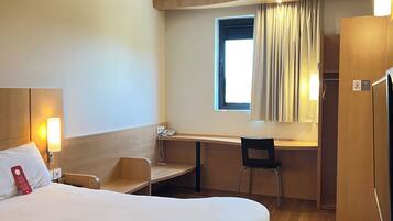 A hotel room with a bed, desk, chair, and a small window with curtains.