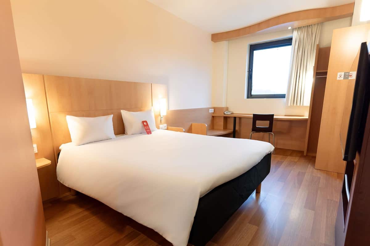 A hotel room with a large bed, a desk, a chair, and a wardrobe.