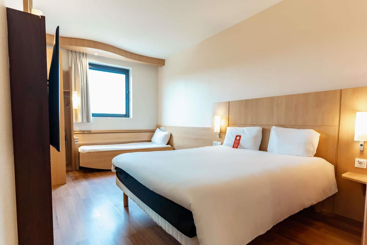 A hotel room with a large bed, a smaller sofa, a window with curtains, a wooden headboard, and a bedside table with a lamp.
