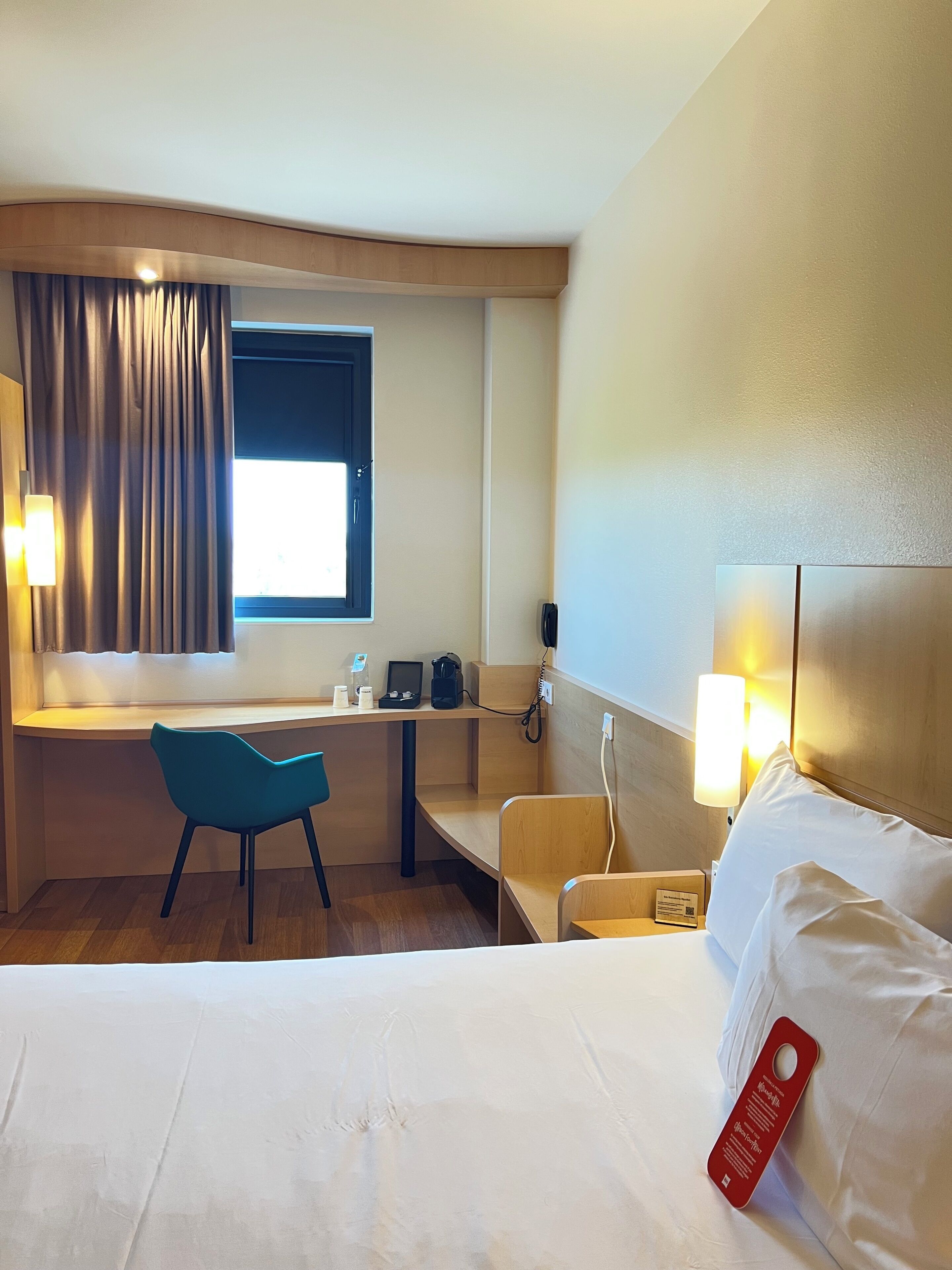 A hotel room with a bed, a desk with a chair, a window with curtains, a bedside table with a lamp, and a bedside phone.