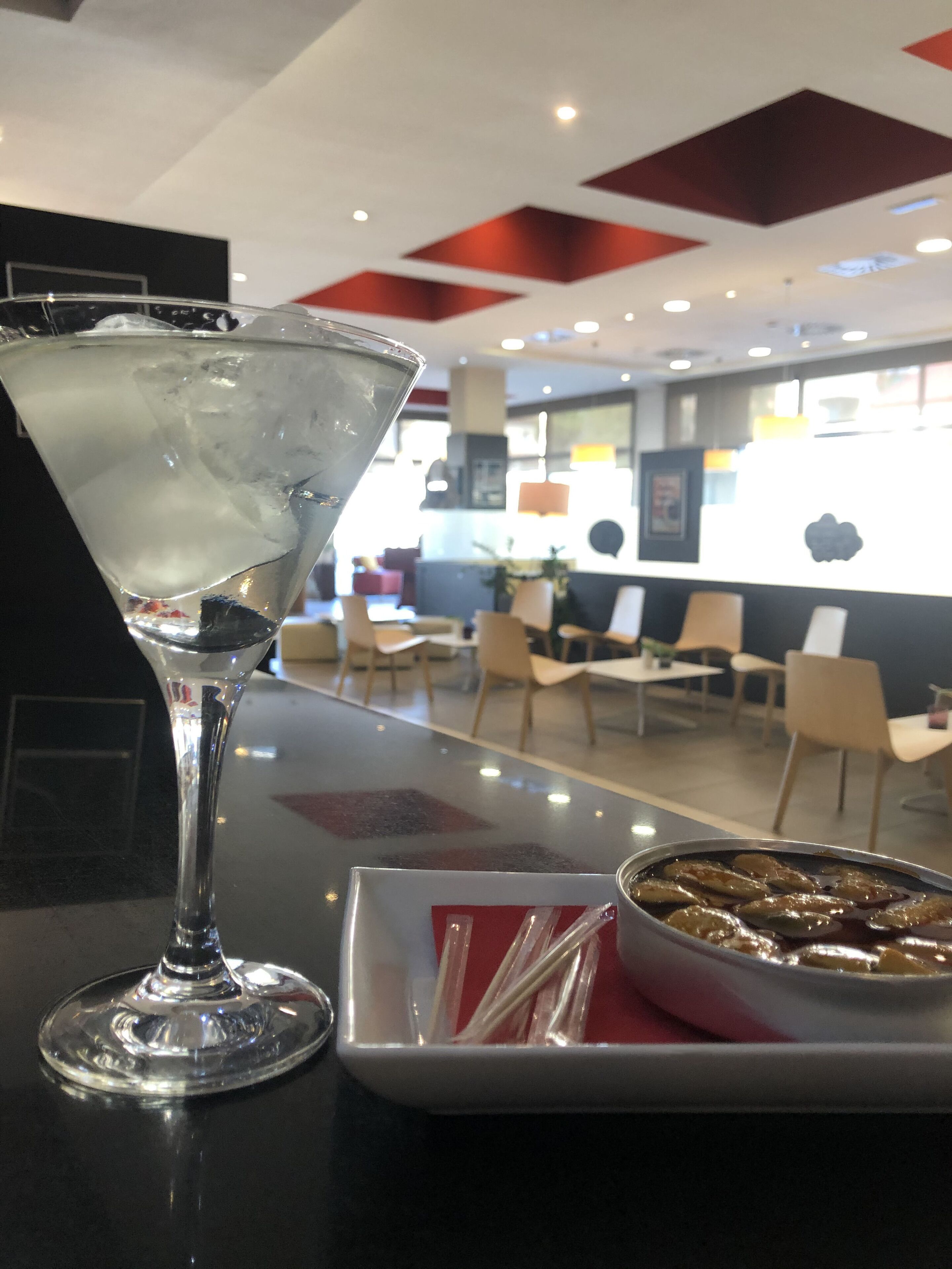 Una copa de martini con hielo sobre una mesa, un plato pequeño con comida y un mantel rojo. El fondo muestra un interior moderno con sillas y mesas.