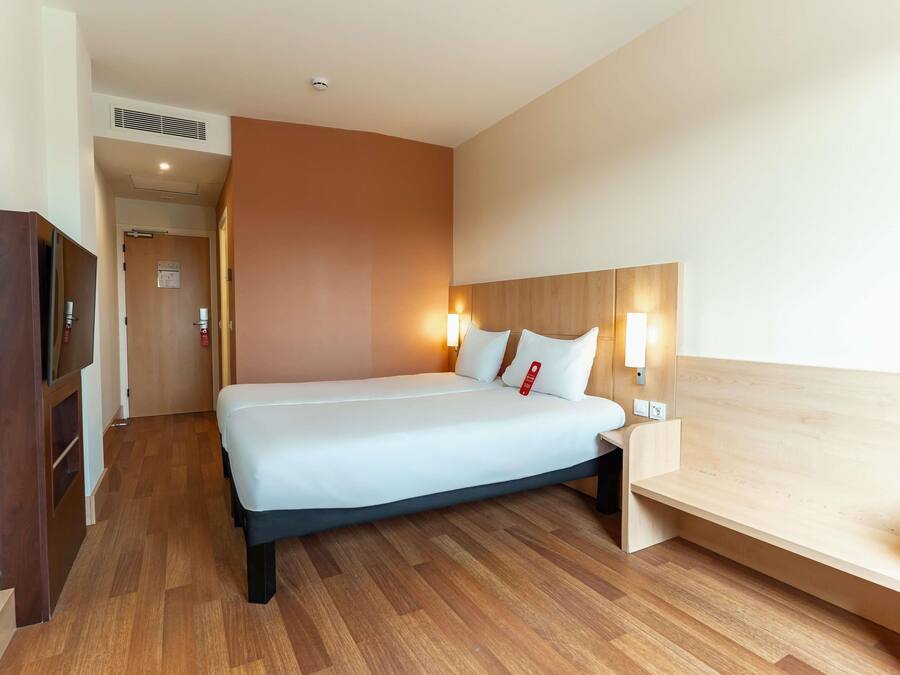 Habitación de hotel con cama, lámparas de noche, cabecero de madera, escritorio de madera y puerta con extintor.