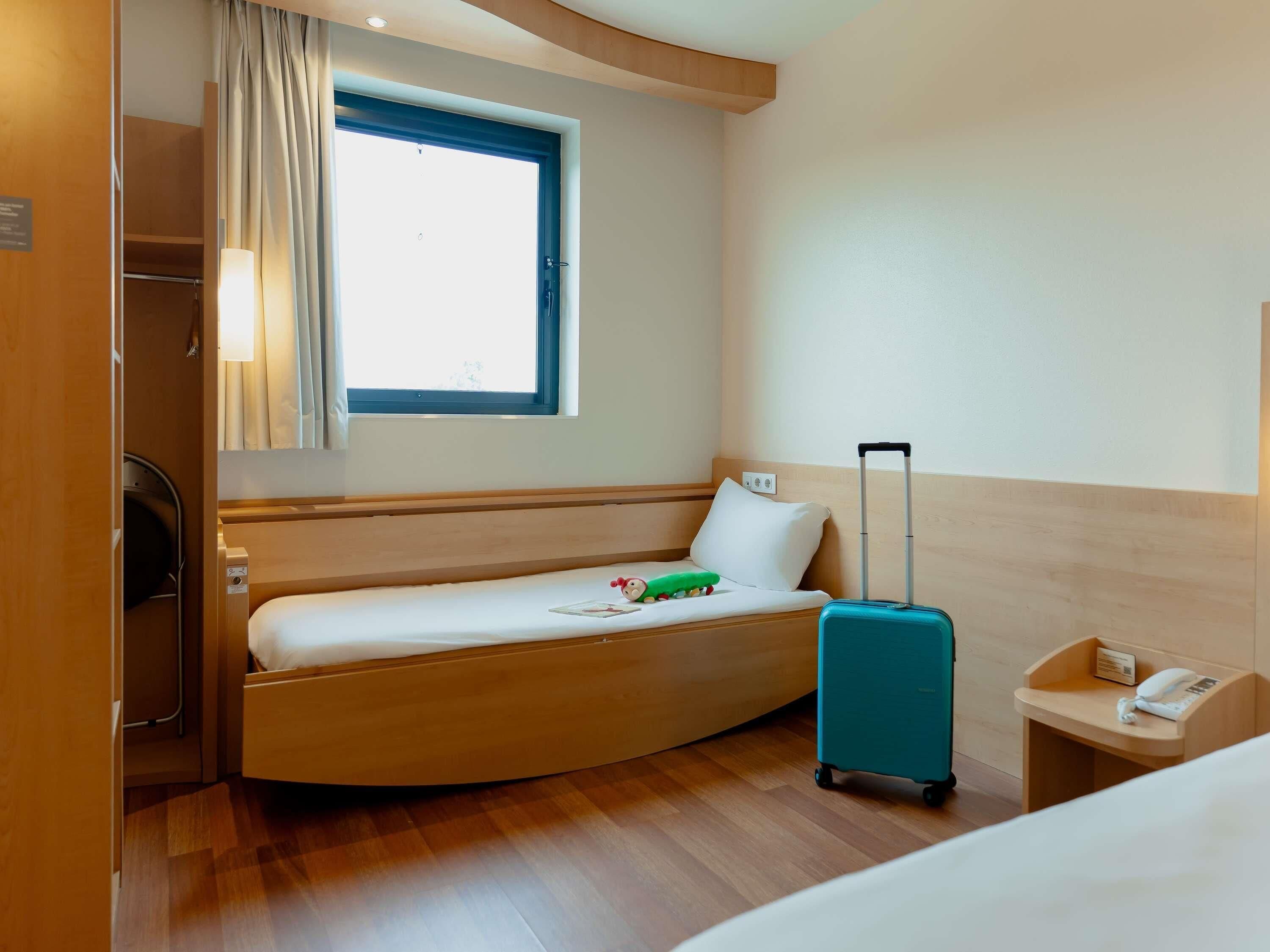 Habitación de hotel pequeña con una cama individual, ventana con cortinas, mesita de noche con un teléfono y una maleta.