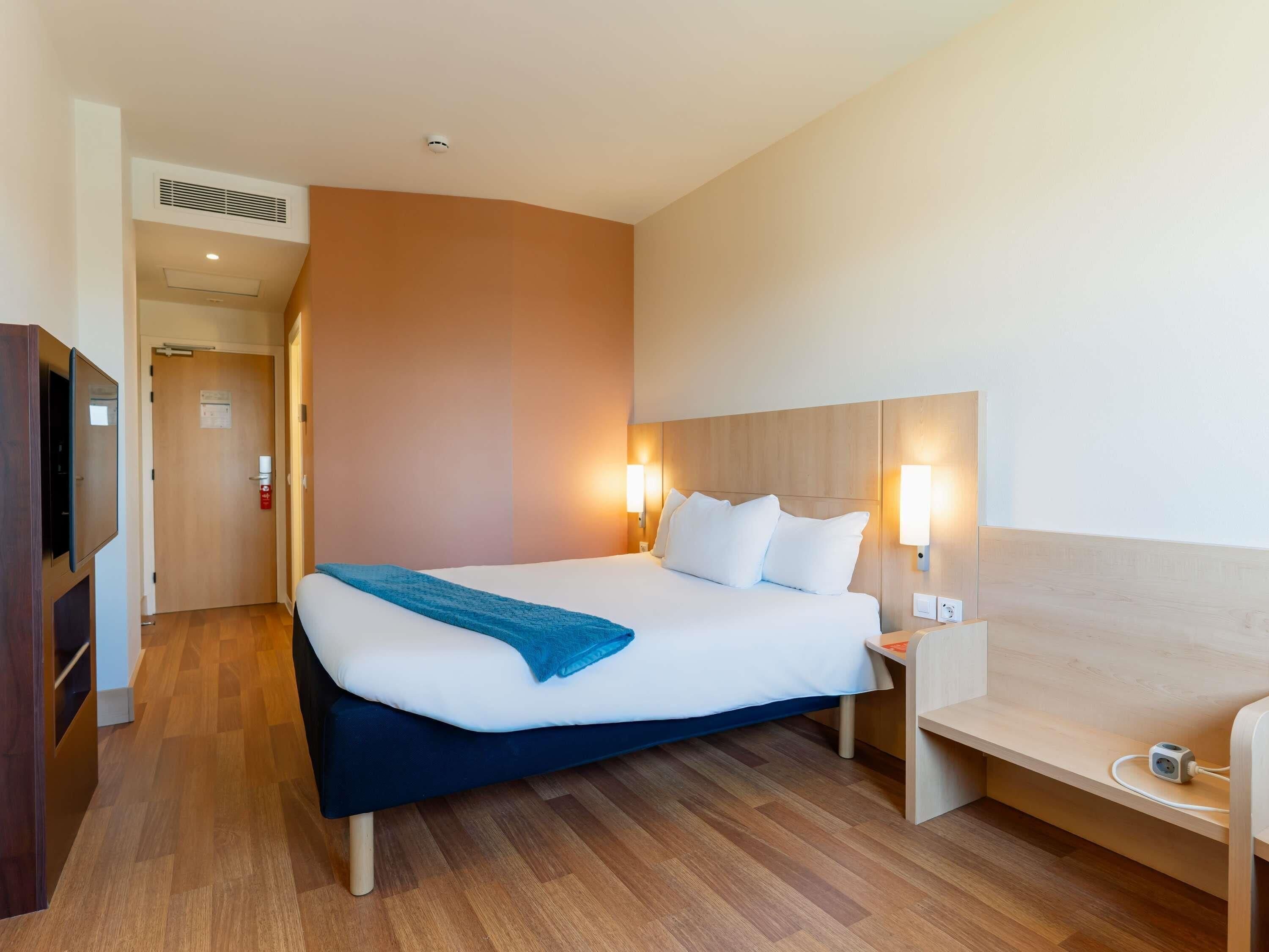 A hotel room with a bed, bedside tables, a desk, a chair, and a TV.