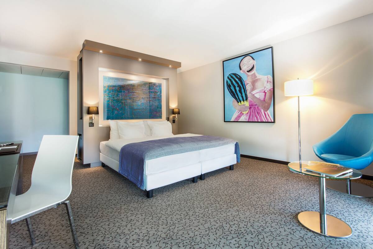Una habitación de hotel moderna con una cama grande, un escritorio, una silla azul y un cuadro en la pared.