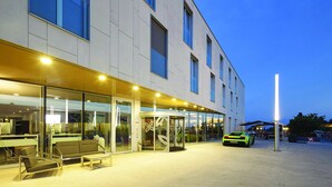 A modern building with a glass entrance, a green car parked outside, and a seating area with a table and chairs. - Starling Hotel Lausanne (Saint-Sulpice)