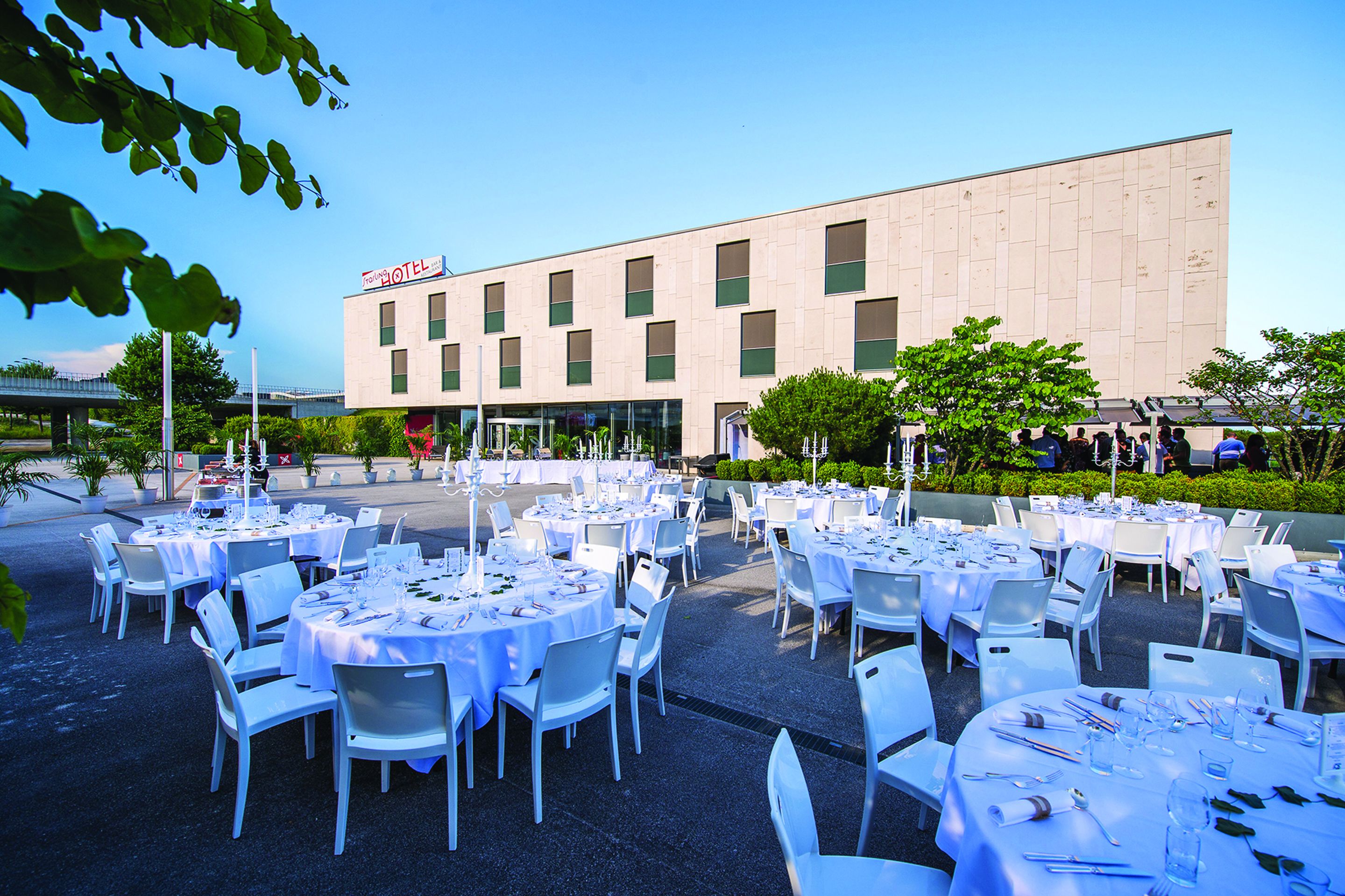 Montaje de banquete al aire libre con mesas redondas y sillas blancas frente a un edificio con un letrero que dice 'Hotel'.